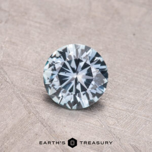 A 0.74-carat Montana Sapphire with a round brilliant cut and symmetrical facets is displayed on a textured beige surface.