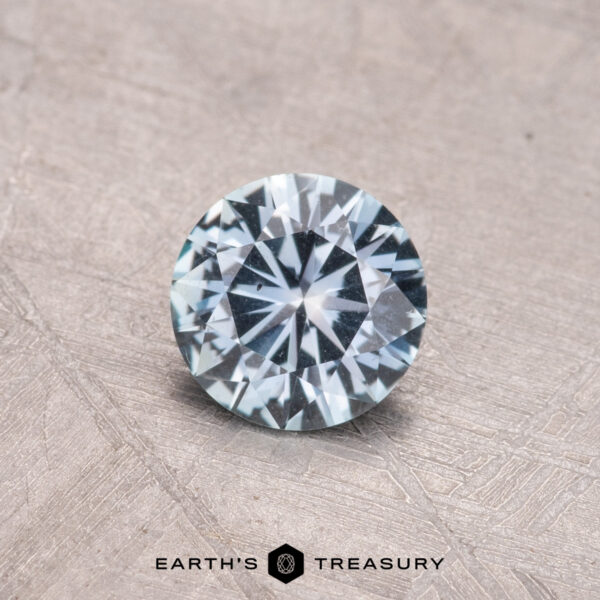 0.74-Carat Montana Sapphire (Heated) A 0.74-carat Montana Sapphire with a round brilliant cut and symmetrical facets is displayed on a textured beige surface.