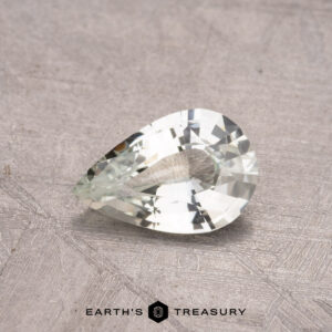 A pear-shaped, near-colorless gemstone weighing 0.87 carats sits on a textured gray surface with "Earth's Treasury" beneath it, echoing the elegance of a rare Montana Sapphire.