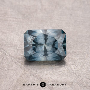 A 1.00-carat faceted rectangular gemstone shines in a deep peacock blue on a textured light gray surface. This heated Montana Sapphire captivates with its rich color and brilliance. At the bottom, the logo reads "Earth's Treasury.