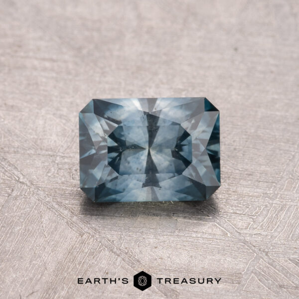 1.00-Carat Montana Sapphire (Heated) A 1.00-carat faceted rectangular gemstone shines in a deep peacock blue on a textured light gray surface. This heated Montana Sapphire captivates with its rich color and brilliance. At the bottom, the logo reads "Earth's Treasury.