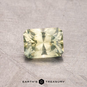 A faceted, rectangular light green 0.98-carat Montana sapphire (heated) is showcased against a textured backdrop. Below the gemstone, the words "Earth's Treasury" are visible.