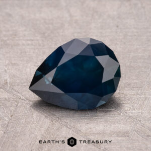 A 1.64-carat pear-shaped Montana sapphire, heated and dark blue with facets, rests on a textured gray surface.
