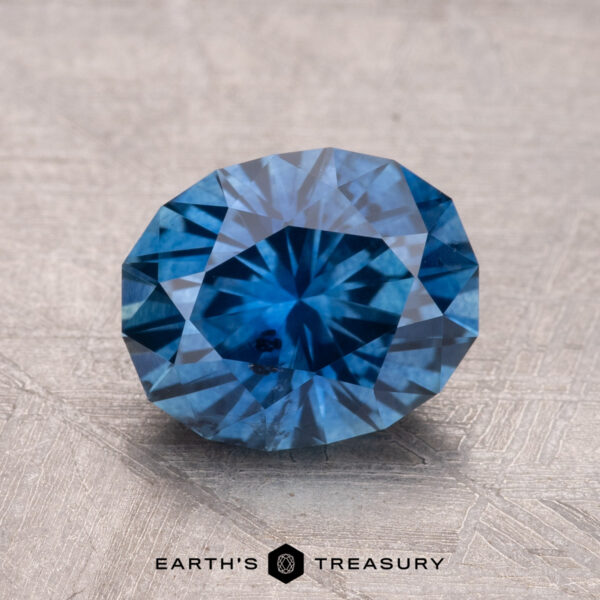 2.43-Carat Montana Sapphire (Heated) A 2.43-carat Montana Sapphire (heated) in a round brilliant-cut displayed on a textured surface with the logo "Earth's Treasury" beneath.