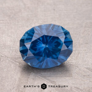 A blue, faceted 2.10-carat Montana sapphire (heated) rests on a textured surface with "Earth's Treasury" written below.
.
