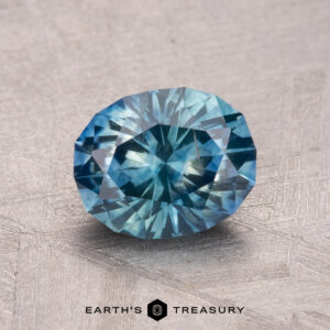 A round, faceted teal gemstone, a 1.81-carat Montana Sapphire (heated), is displayed on a textured surface with the text "Earth's Treasury" beneath it.