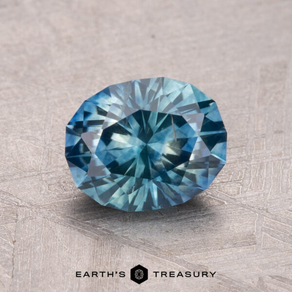 1.81-Carat Montana Sapphire (Heated) A round, faceted teal gemstone, a 1.81-carat Montana Sapphire (heated), is displayed on a textured surface with the text "Earth's Treasury" beneath it.