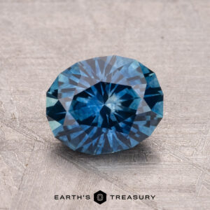 A 1.80-carat, round-cut, faceted blue Montana Sapphire (heated) rests on a textured surface with "Earth's Treasury" displayed at the bottom.