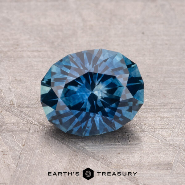 A 1.80-carat, round-cut, faceted blue Montana Sapphire (heated) rests on a textured surface with "Earth's Treasury" displayed at the bottom.