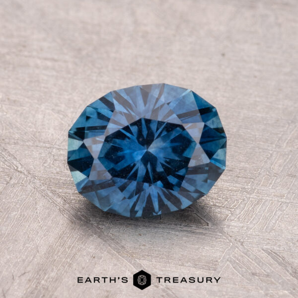 1.79-Carat Montana Sapphire (Heated) A 1.79-carat heated Montana Sapphire, round and blue with facets, rests on a textured surface displaying the "Earth's Treasury" logo.