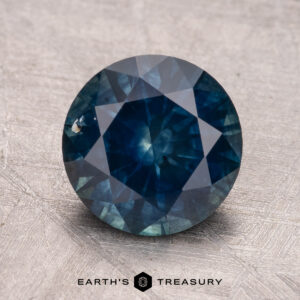 A 2.83-carat Montana Sapphire (heated), round and dark blue with visible facets, set against a textured gray surface.