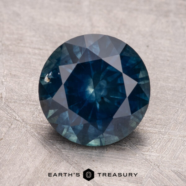 2.83-Carat Montana Sapphire (Heated) A 2.83-carat Montana Sapphire (heated), round and dark blue with visible facets, set against a textured gray surface.