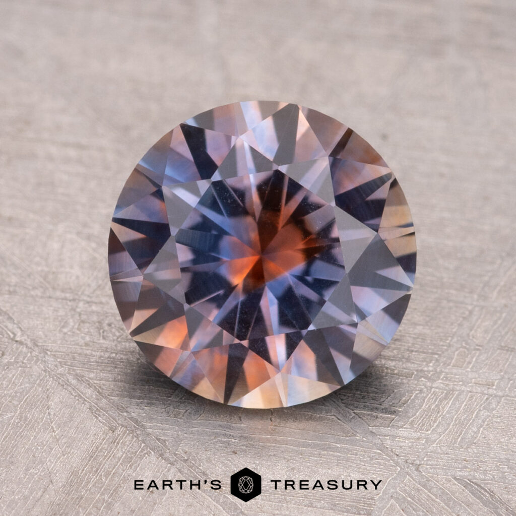 A textured, round faceted gemstone with blue and orange hues, featuring the "Earth's Treasury" logo at the bottom. Product: 2.54-Carat Montana Sapphire (Heated).