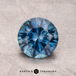 A 1.68-carat Montana sapphire (heated) with a round brilliant cut and intricate facets, displayed on a textured surface.