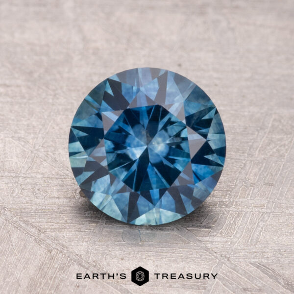 1.68-Carat Montana Sapphire (Heated) A 1.68-carat Montana sapphire (heated) with a round brilliant cut and intricate facets, displayed on a textured surface.