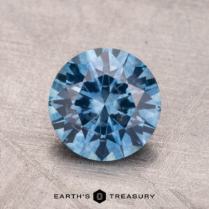 A 1.66-carat Montana sapphire (heated) with a round brilliant cut rests on a textured surface.