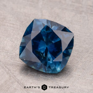 A 2.40-carat cushion-cut Montana sapphire with a blue hue is showcased on a textured surface featuring the "Earth's Treasury" logo.