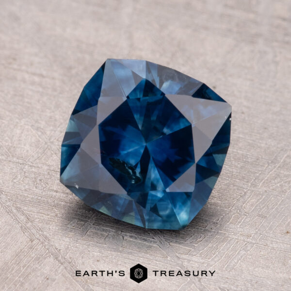 2.40-Carat Montana Sapphire (Heated) A 2.40-carat cushion-cut Montana sapphire with a blue hue is showcased on a textured surface featuring the "Earth's Treasury" logo.