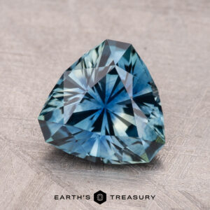 A 2.26-carat Montana sapphire, heart-shaped and blue-green with intricate facets, sits on a textured surface.