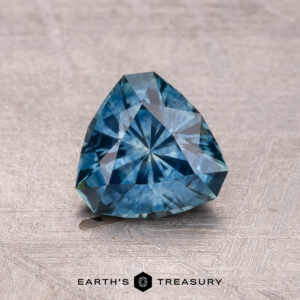 A trillion-cut bluish gemstone with a faceted surface rests on a textured background, featuring the "Earth's Treasury" logo at the bottom. It is a 1.17-carat heated Montana Sapphire.