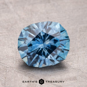 A faceted 2.30-carat heated Montana sapphire with a starburst pattern rests on a textured surface. Text at the bottom reads "Earth's Treasury.