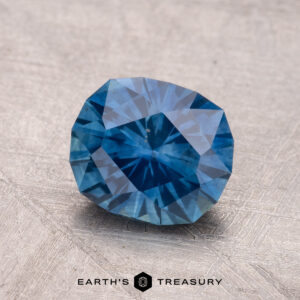 A 1.91-carat heated Montana sapphire, faceted and blue, rests on a textured surface with the "Earth's Treasury" logo beneath it.
