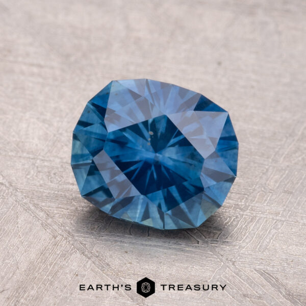 1.91-Carat Montana Sapphire (Heated) A 1.91-carat heated Montana sapphire, faceted and blue, rests on a textured surface with the "Earth's Treasury" logo beneath it.