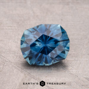 A faceted blue 1.82-carat heated Montana Sapphire with a geometric pattern rests on a textured surface, labeled "Earth's Treasury" at the bottom.
