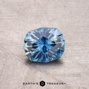 A 0.80-carat heated Montana sapphire with a faceted cut is showcased on a textured gray surface, with "Earth's Treasury" visible beneath.