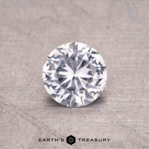 A round, faceted 0.72-carat Montana sapphire rests on a textured surface with "Earth's Treasury" printed below.
