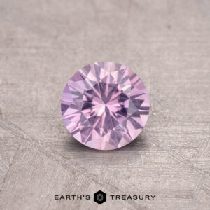 The 0.68-carat Montana Sapphire, a round, faceted pink gemstone, is displayed against a textured surface.