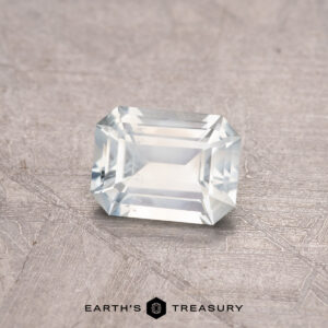 A rectangular, faceted white gemstone on a textured gray surface with "0.82-Carat Montana Sapphire" written below.