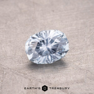 A sparkling oval-cut gemstone with a light blue hue, reminiscent of a nearly colorless 0.75-Carat Montana Sapphire, rests on a textured surface. Text reads "Earth's Treasury.