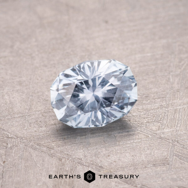 0.75-Carat Montana Sapphire A sparkling oval-cut gemstone with a light blue hue, reminiscent of a nearly colorless 0.75-Carat Montana Sapphire, rests on a textured surface. Text reads "Earth's Treasury.
