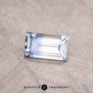 A 0.50-carat Montana sapphire gleams in a rectangular cut, highlighting its mirrored effect with blue, white, and pale lavender tones on a textured surface.