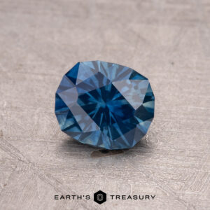 A 1.01-carat heated Montana sapphire gemstone, round and teal, on a textured surface.