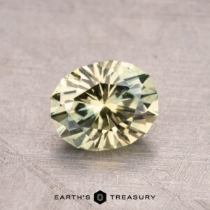 A faceted pale yellow 1.32-carat Heated Montana Sapphire rests on a textured surface, with the Earth's Treasury logo beneath it.