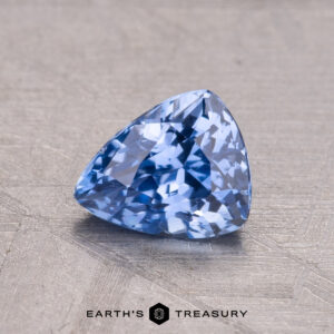 A 1.32-carat trillion-cut blue Ceylon Sapphire rests on a textured surface, with "Earth's Treasury" visible below.