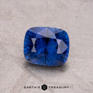 A 1.32-carat Ceylon sapphire, heated and geometric cut, rests on a textured gray surface with "Earth's Treasury" displayed at the bottom.