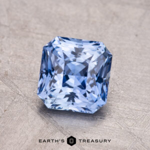 The 1.54-Carat Ceylon Sapphire, a faceted rectangular blue gemstone with a sparkling surface, is displayed on a textured gray background.