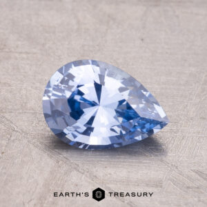 A pear-shaped, faceted blue gemstone rests on a textured surface with "1.44-Carat Ceylon Sapphire" labeled below.