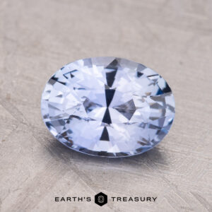 A faceted oval 2.23-Carat Ceylon Sapphire gemstone with a light blue hue is displayed against a textured background, with "Earth's Treasury" at the bottom.