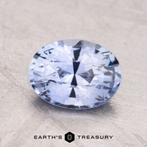 A faceted oval 2.23-Carat Ceylon Sapphire gemstone with a light blue hue is displayed against a textured background, with "Earth's Treasury" at the bottom.
