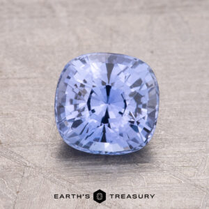 A 1.55-carat Ceylon sapphire in a cushion cut sits on a textured surface with the "Earth's Treasury" logo at the bottom.