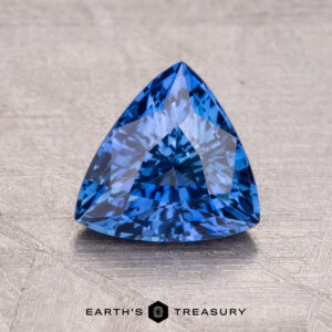 A 1.52-carat Ceylon Sapphire, triangular and intricately faceted, rests on a textured surface.