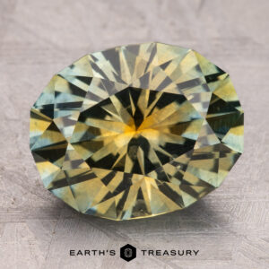 An 8.75-carat, faceted round Montana Sapphire in teal and yellow rests on a textured surface, labeled "Earth's Treasury.