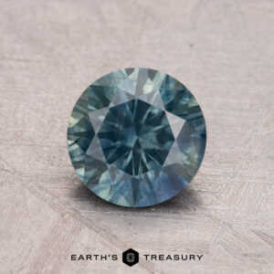 A round faceted blue-green 1.50-carat Montana Sapphire (heated) rests on a textured surface with the words "Earth's Treasury" below.
