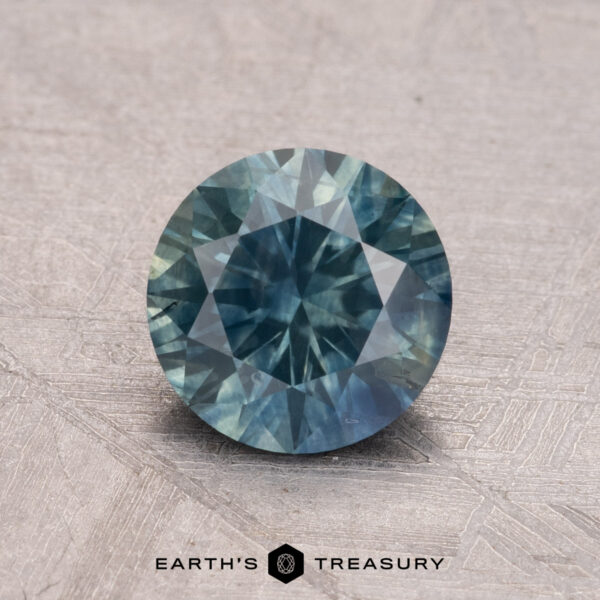 1.50-Carat Montana Sapphire (Heated) A round faceted blue-green 1.50-carat Montana Sapphire (heated) rests on a textured surface with the words "Earth's Treasury" below.