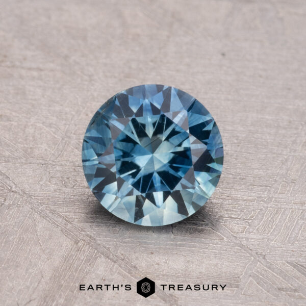 1.15-Carat Montana Sapphire (Heated) A 1.15-carat heated Montana sapphire, round and blue, is placed on a textured surface with the "Earth's Treasury" logo below.