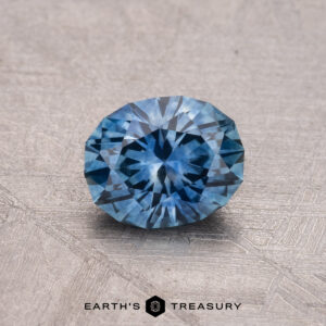 A round 0.98-carat heated Montana sapphire with intricate facets rests on a textured surface. Text reads: Earth’s Treasury.
